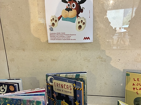 In der Bibliothek stehen Kinderbücher unter einem Poster mit einem Maskottchen