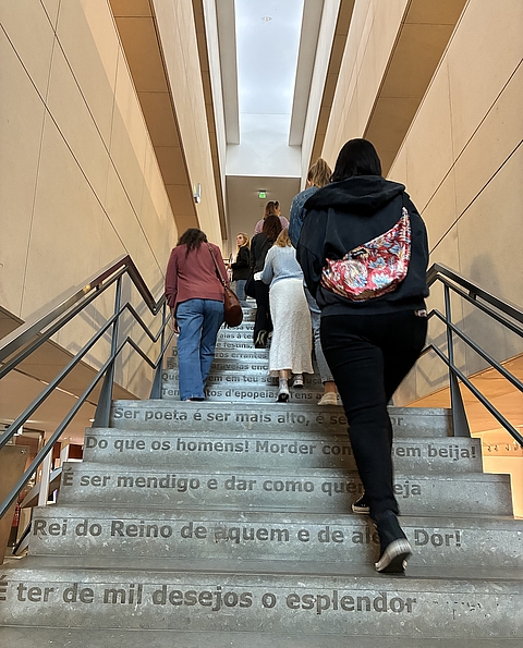 Die Gruppe steigt eine Treppe in der Bibliothek hoch. Auf jeder Stufe ist eine Zeile portugiesischer Text.